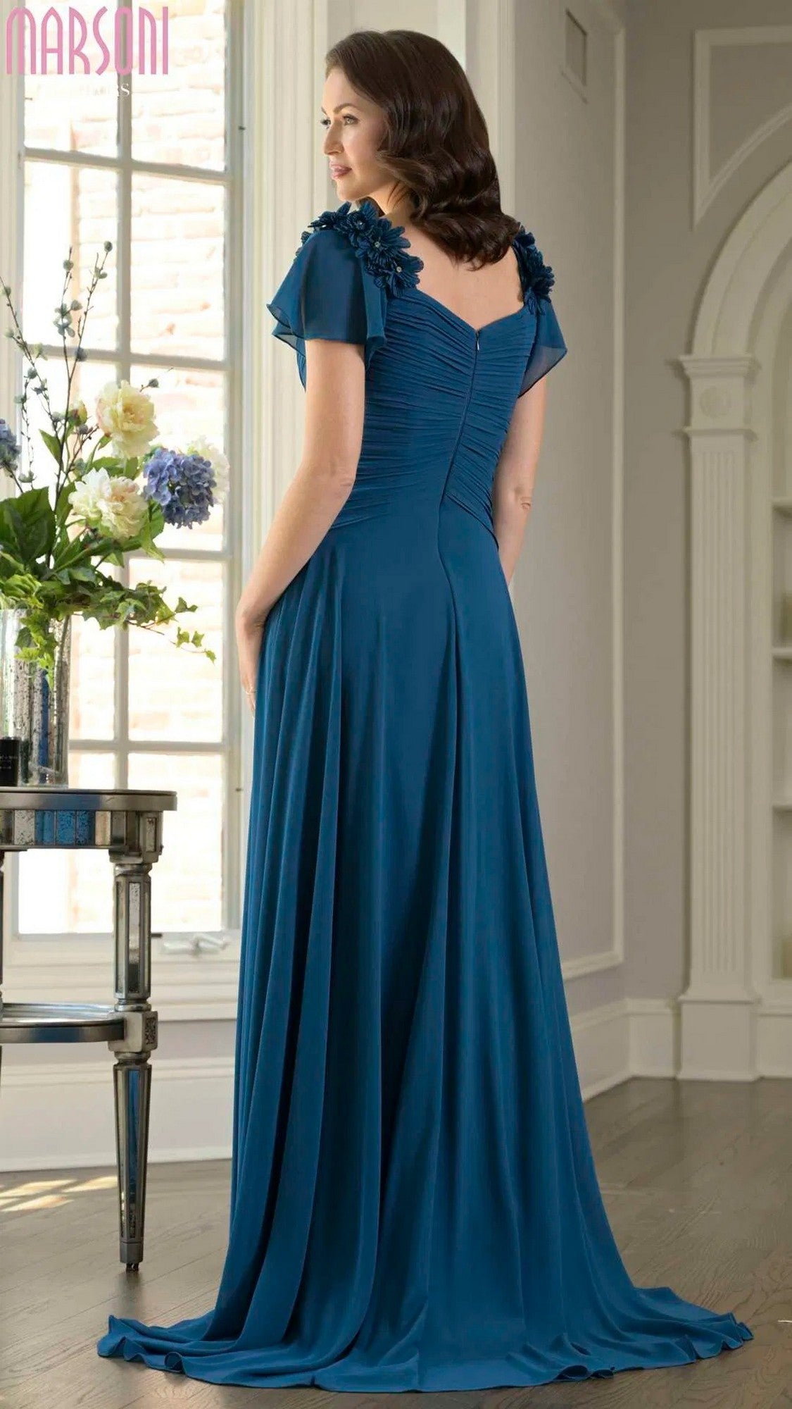 A woman stands indoors facing away, wearing the Marsoni M338 long formal dress in teal chiffon with 3D floral appliques on the sleeves and an elegant A-line skirt.
